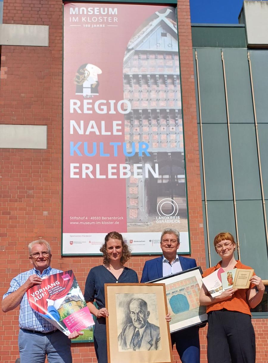 Vor dem acht Meter hohen Banner am Kreishaus werden Objekte rund um das Jubiläum des Bersenbrücker Museums präsentiert (von links): Kulturbüroleiter Burkhard Fromme mit einem Plakat für das Theater „Vorhang auf!“, Museumsleiterin Katharina Pfaff mit einem Portrait von Museumsgründer Rothert, Kreisrat Matthias Selle mit einem Aquarell Theodor Doebners und Museumspädagogin Claudia Stuckenberg mit Museumsführern aus drei Epochen. Foto: Landkreis Osnabrück/Henning Müller-Detert