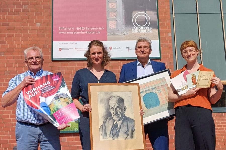 Vor dem acht Meter hohen Banner am Kreishaus werden Objekte rund um das Jubiläum des Bersenbrücker Museums präsentiert (von links): Kulturbüroleiter Burkhard Fromme mit einem Plakat für das Theater „Vorhang auf!“, Museumsleiterin Katharina Pfaff mit einem Portrait von Museumsgründer Rothert, Kreisrat Matthias Selle mit einem Aquarell Theodor Doebners und Museumspädagogin Claudia Stuckenberg mit Museumsführern aus drei Epochen. Foto: Landkreis Osnabrück/Henning Müller-Detert