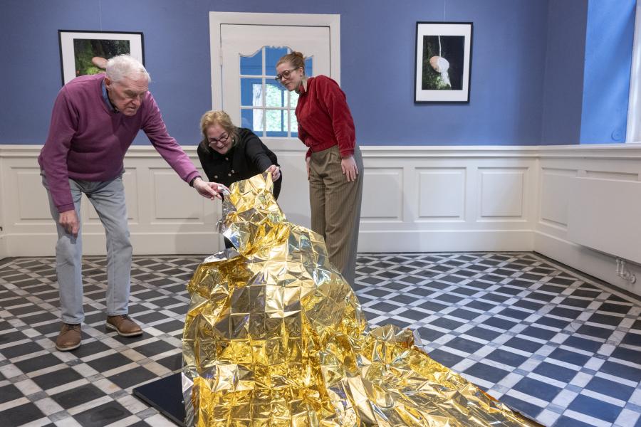 Zu sehen ist der Blaue Salon des Museums im Klosters. Im Vordergrund befindet sich ein mit einer goldenen Decke abgedeckten Objekt. Unter diese Decke schauen dahinterstehend drei Personen: Vorsitzender des Kreisheimatbundes Bersenbrück, Künstlerin Sigrun Menzel und Museumsleitung Kim Winkelmann